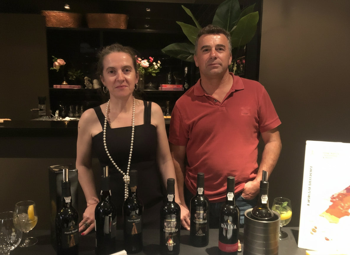 Two people standing behind a table with wine bottles and glasses in a dimly lit room.